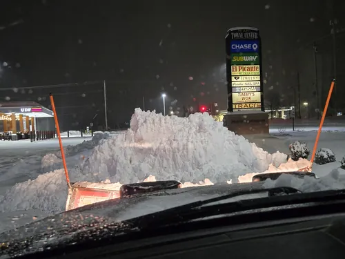 View over a plow blade pushing snow into a pile at a lit commercial plaza at night